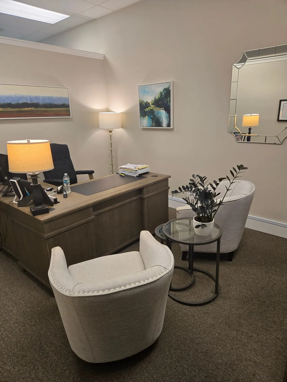 A private office with a lamp, landscape art, wood desk, and two cream guest chairs